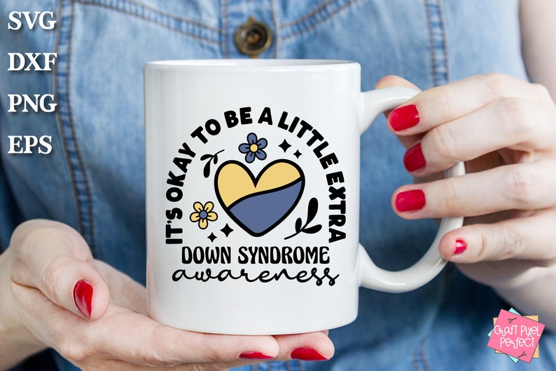May include: White ceramic mug with a black and blue design. The design features a heart with yellow and blue colors, flowers, and the text "It's okay to be a little extra Down Syndrome awareness".