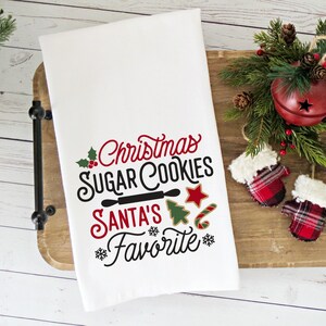 May include: White kitchen towel with the text "Christmas Sugar Cookies Santa's Favorite" in red and black lettering. The text is surrounded by a holly sprig, a rolling pin, a star, a candy cane, and a Christmas tree.