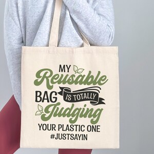 Pu&ograve; includere: Una borsa di tela bianca con il testo "My Reusable Bag is Totally Judging Your Plastic One #JUSTSAYIN" in lettere nere e verdi. La borsa &egrave; tenuta da una persona che indossa un maglione grigio e pantaloni marroni.