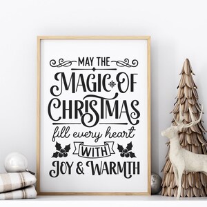 Puede incluir: Una impresi&oacute;n enmarcada blanca con texto negro que dice "Que la magia de la Navidad llene cada coraz&oacute;n de alegr&iacute;a y calidez". La impresi&oacute;n se muestra en un estante blanco con dos &aacute;rboles decorativos y una figura de ciervo.