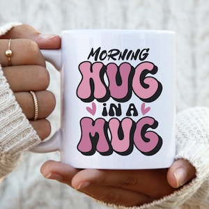 Puede incluir: Taza de cerámica blanca con el texto "Morning Hug in a Mug" en letras rosa y negras. El texto está en una fuente retro, con forma de burbuja.