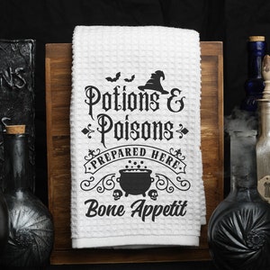 Pu&ograve; includere: Un asciugamano da cucina bianco con un design in bianco e nero che dice "Pozioni e Veleni Preparati Qui. Buon appetito". Il design include un cappello da strega, pipistrelli e un calderone.