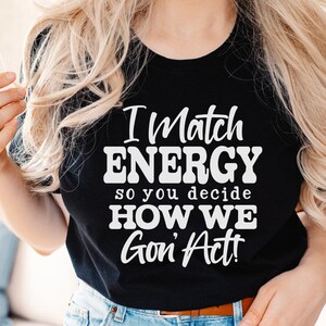 May include: Black t-shirt with the text "I Match ENERGY so you decide HOW WE Gon' Act!" in white. The shirt also has SVG, DXF, PNG, and EPS text on the left side. The shirt is worn by a person with blonde hair.