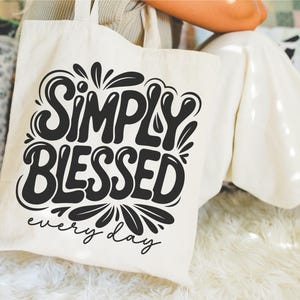 May include: Cream-colored tote bag with the words "SIMPLY BLESSED" in a bold, black, stylized font. Below the main text, the phrase "every day" is written in a smaller, cursive font. The bag is held by a person.