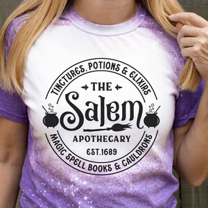 Pu&ograve; includere: Una maglietta bianca con un design circolare nero che dice "The Salem Apothecary Est. 1689" con le parole "Tinctures, Potions & Elixirs" e "Magic Spell Books & Cauldrons" intorno al cerchio.