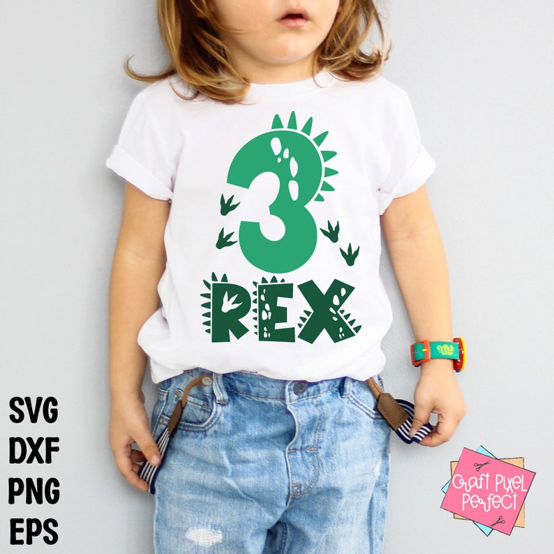 Op de afbeelding: Een wit t-shirt met een groen dinosaurusontwerp. Het nummer "3" staat prominent afgebeeld, samen met het woord "REX" en dinosaurusvoetstappen. Het shirt wordt gedragen door een kind, gecombineerd met een spijkerbroek en een kleurrijk horloge.