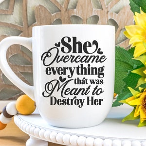 May include: White ceramic mug with a black graphic that reads "She Overcame everything that was Meant to Destroy Her". The mug is sitting on a white pedestal with a string of beads in the foreground.