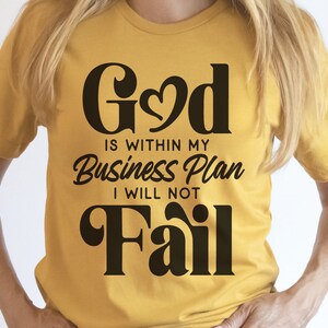 May include: A yellow t-shirt with the text "God is within my Business Plan I will not Fail" in black lettering. The word "God" has a heart in the middle of the "o".