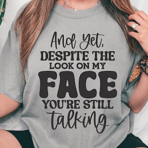 May include: A gray t-shirt with the text "And yet, DESPITE THE LOOK ON MY FACE YOU'RE STILL talking" printed in black.