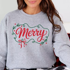 May include: A light gray sweatshirt with the word "Merry" in red script, framed by a green wreath with red berries and a bow. The sweatshirt is a crew neck style, perfect for the holiday season.