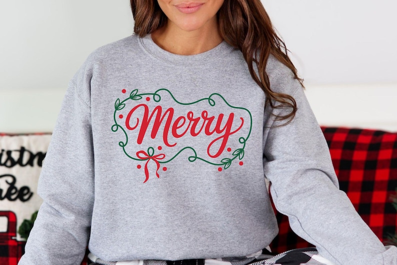 May include: A light gray sweatshirt with the word "Merry" in red script, framed by a green wreath with red berries and a bow. The sweatshirt is a crew neck style, perfect for the holiday season.