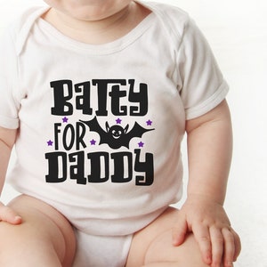 K&ouml;nnte beinhalten: Ein wei&szlig;er Baby-Strampler mit einem schwarzen Aufdruck, der "Batey for Daddy" mit einer schwarzen Fledermaus und lila Sternen sagt.