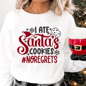 May include: White sweatshirt with red and black text that reads "I ate Santa's cookies #noregrets". The text is surrounded by snowflakes and a red Santa hat. A person is wearing the sweatshirt and holding a red and black Santa mug.