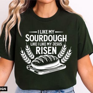 May include: A green t-shirt with a white graphic design that says "I like my sourdough like I like my Jesus risen". The graphic also includes a loaf of bread on a plate and wheat stalks.