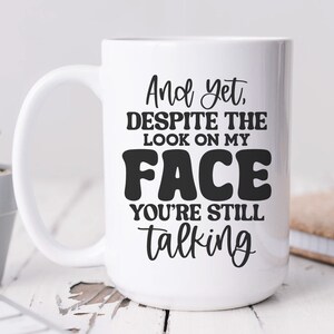 May include: White ceramic coffee mug with black text that reads "And yet, despite the look on my FACE you're still talking".