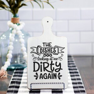 May include: A white wooden cutting board with a handle, featuring the text "THE DISHES looking at me DIRTY AGAIN" in black. The board is displayed on a black and white checkered cloth.