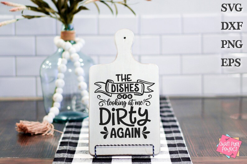 May include: A white wooden cutting board with a handle, featuring the text "THE DISHES looking at me DIRTY AGAIN" in black. The board is displayed on a black and white checkered cloth.