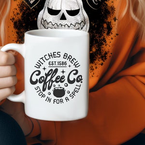 以下が含まれることがあります： 「Witches Brew Coffee Co. Est. 1586 Stop in for a Spell」と書いてある白地に黒と白のグラフィックデザインが施された白いコーヒーカップ。グラフィックには泡が立った釜が描かれている