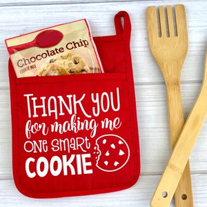 May include: Red oven mitt with a white pocket and the text "THANK YOU for making me ONE SMART COOKIE" with a cookie graphic.  The oven mitt is on a white wood surface with a bag of chocolate chip cookie mix and two wooden kitchen utensils.