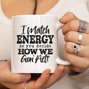 May include: White ceramic mug with the text "I Match ENERGY so you decide HOW WE Gon' Act!" in black. The mug is held by a person wearing multiple rings. The mug is a standard size for coffee or tea.