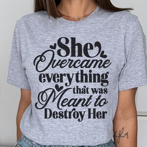 May include: A gray t-shirt with the text "She Overcame everything that was Meant to Destroy Her" printed in black.