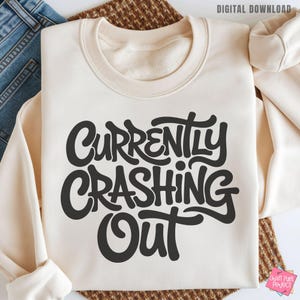 May include: Cream-colored sweatshirt with the phrase "Currently Crashing Out" in bold, black, stylized lettering. The sweatshirt is folded, with the sleeves visible. The words "Digital Download" are in the top right corner.