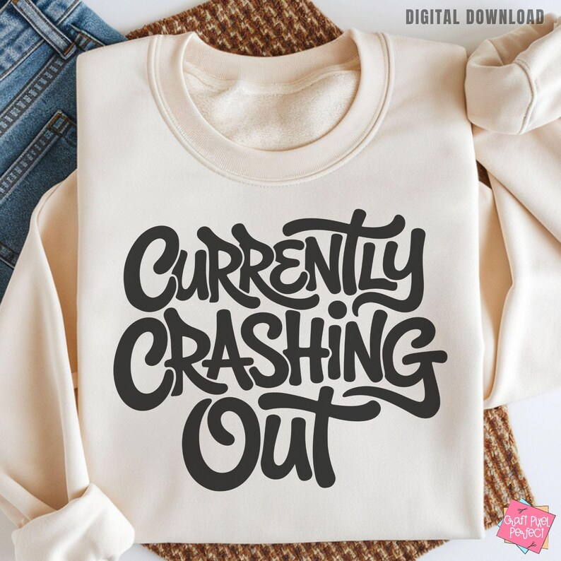 May include: Cream-colored sweatshirt with the phrase "Currently Crashing Out" in bold, black, stylized lettering. The sweatshirt is folded, with the sleeves visible. The words "Digital Download" are in the top right corner.