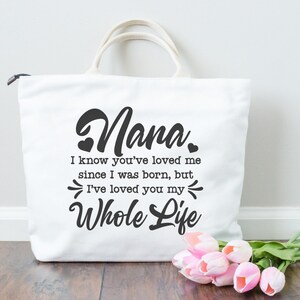 May include: White tote bag with black text that reads "Nana. I know you've loved me since I was born, but I've loved you my whole life."  The bag is sitting on a wooden surface with a bouquet of pink flowers.