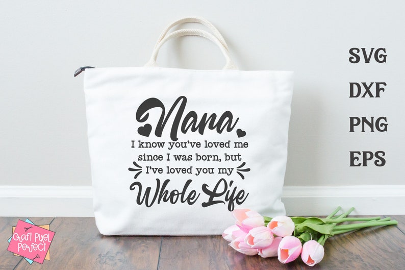 May include: White tote bag with black text that reads "Nana. I know you've loved me since I was born, but I've loved you my whole life."  The bag is sitting on a wooden surface with a bouquet of pink flowers.