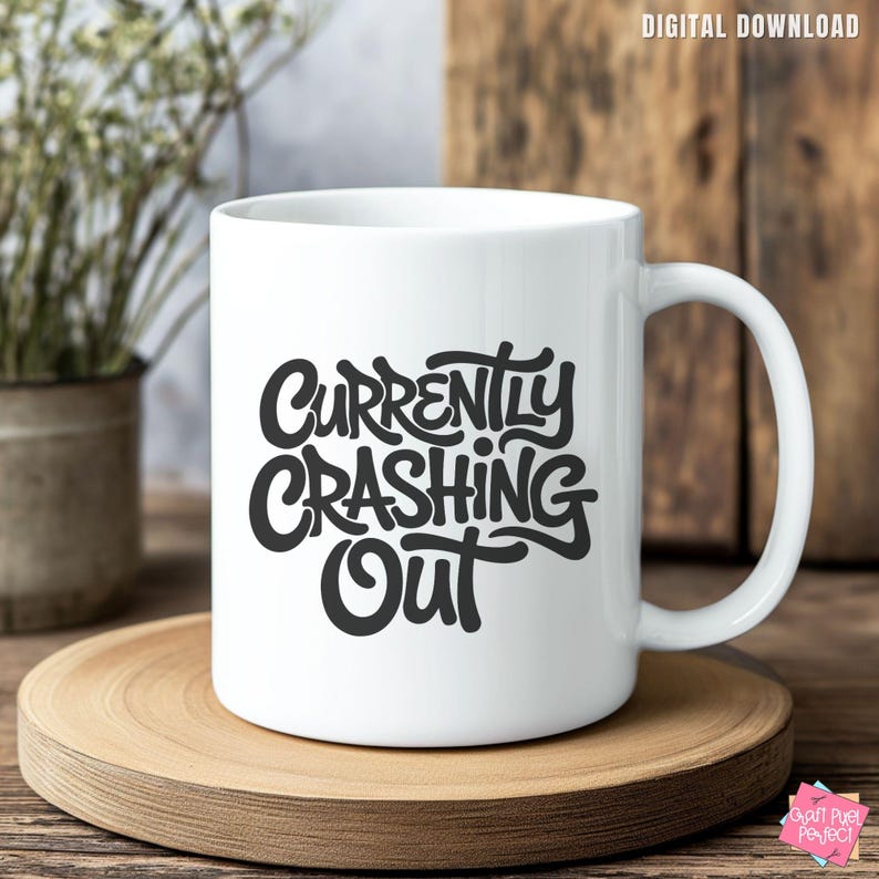 May include: White ceramic mug with the words "Currently Crashing Out" in black script. The mug has a curved handle and is sitting on a wooden coaster. The mug is a digital download.