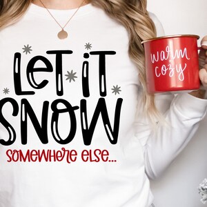 May include: White long-sleeved shirt with black text that reads "Let it snow somewhere else..." A red mug with white text that reads "warm cozy" is being held by a person wearing the shirt.