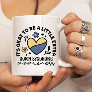 May include: White ceramic mug with a blue and yellow heart and the text "It's okay to be a little extra Down Syndrome awareness".