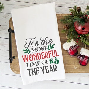 May include: White kitchen towel with the text "It's the MOST WONDERFUL TIME OF THE YEAR" in red and gray lettering with green sprigs.