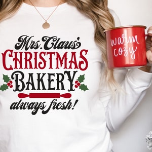K&ouml;nnte beinhalten: Ein wei&szlig;es Langarmshirt mit dem Text "Mrs. Claus's Christmas Bakery always fresh!" in schwarzer und roter Schrift. Das Shirt zeigt einen roten Nudelholz und Stechpalmenbl&auml;tter mit roten Beeren.