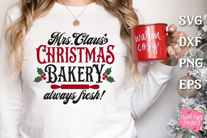 K&ouml;nnte beinhalten: Ein wei&szlig;es Langarmshirt mit dem Text "Mrs. Claus's Christmas Bakery always fresh!" in schwarzer und roter Schrift. Das Shirt zeigt einen roten Nudelholz und Stechpalmenbl&auml;tter mit roten Beeren.