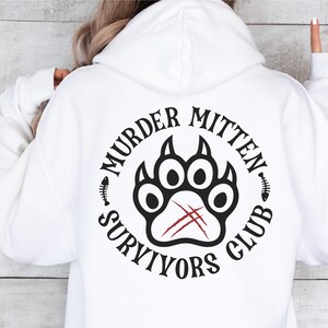 Pu&ograve; includere: Felpa bianca con un design grafico nero e rosso che dice "Murder Mitten Survivors Club" con un'impronta di zampa al centro.