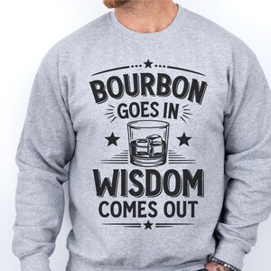May include: A gray sweatshirt with the text "Bourbon goes in Wisdom comes out" printed on the front. The text is surrounded by stars and a graphic of a glass of bourbon.