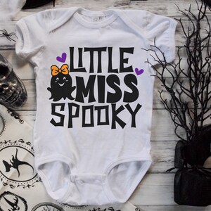 K&ouml;nnte beinhalten: Wei&szlig;er Baby-Strampler mit einem schwarzen Geistermotiv und dem Text "Little Miss Spooky" in Schwarz mit violetten Akzenten.