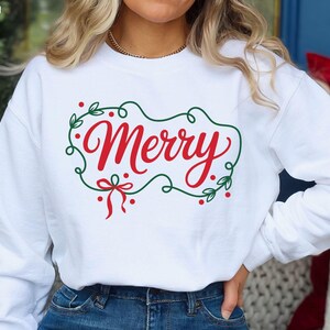 May include: White sweatshirt with the word "Merry" in red script, framed by a green wreath with red berries and a bow. The design is a digital graphic, perfect for holiday apparel.