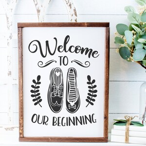 May include: A white wooden sign with a brown frame, featuring the text "Welcome to Our Beginning" in black cursive font. The sign also includes a pair of black shoes with a heart detail and two small black leaf sprigs.