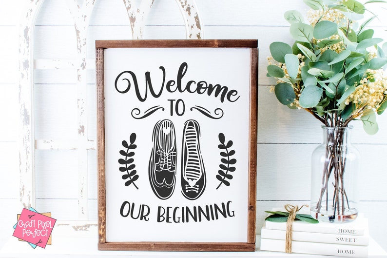 May include: A white wooden sign with a brown frame, featuring the text "Welcome to Our Beginning" in black cursive font. The sign also includes a pair of black shoes with a heart detail and two small black leaf sprigs.