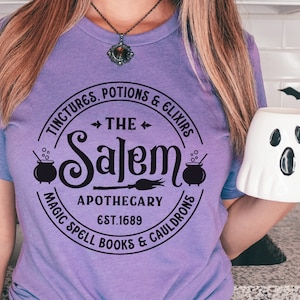 Pu&ograve; includere: Maglietta viola con un design circolare nero che dice "The Salem Apothecary Est. 1689" con immagini di calderoni e una scopa.