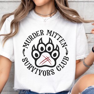 Pu&ograve; includere: Maglietta bianca con un design grafico nero e rosso. La grafica presenta un'impronta di zampa con il testo "Murder Mitten Survivors Club".
