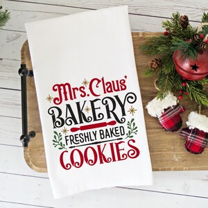 May include: White kitchen towel with red and black text that reads "Mrs. Claus' Bakery Freshly Baked Cookies". The towel is on a wooden tray with a red bell and red and black mittens.