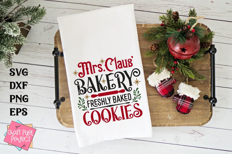 May include: White kitchen towel with red and black text that reads "Mrs. Claus' Bakery Freshly Baked Cookies". The towel is on a wooden tray with a red bell and red and black mittens.