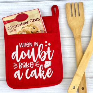May include: Red oven mitt with a white pocket and the text "When in doubt bake a cake" with a pie slice graphic. The oven mitt is hanging on a white wooden surface with two wooden spoons.