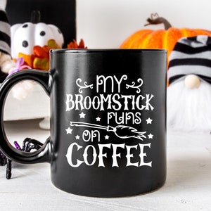 Peut inclure: Une tasse &agrave; caf&eacute; noire avec un motif blanc qui dit "My broomstick runs on coffee" avec un petit balai et des &eacute;toiles. La tasse est pos&eacute;e sur une surface blanche avec une citrouille, des gnomes et des araign&eacute;es en arri&egrave;re-plan.