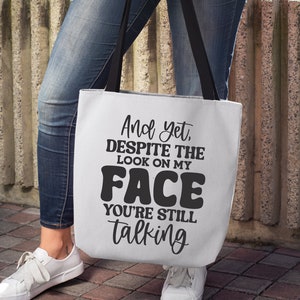 May include: A white tote bag with black text that reads "And yet, despite the look on my face you're still talking".