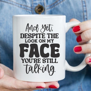 May include: White ceramic mug with black text that reads "And yet, despite the look on my face you're still talking".