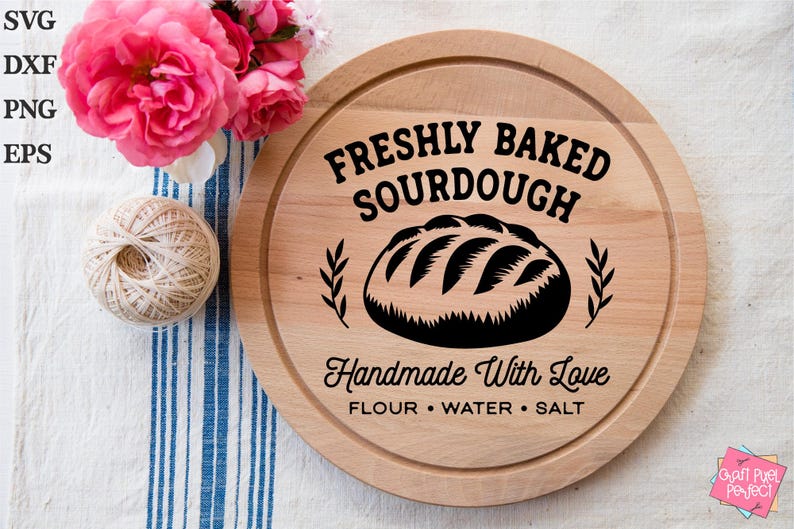 May include: A circular wooden chopping board with the text "FRESHLY BAKED SOURDOUGH" and "Handmade With Love". The board features a bread illustration and is surrounded by pink roses and a ball of twine. The text also includes "FLOUR &bull; WATER &bull; SALT".
